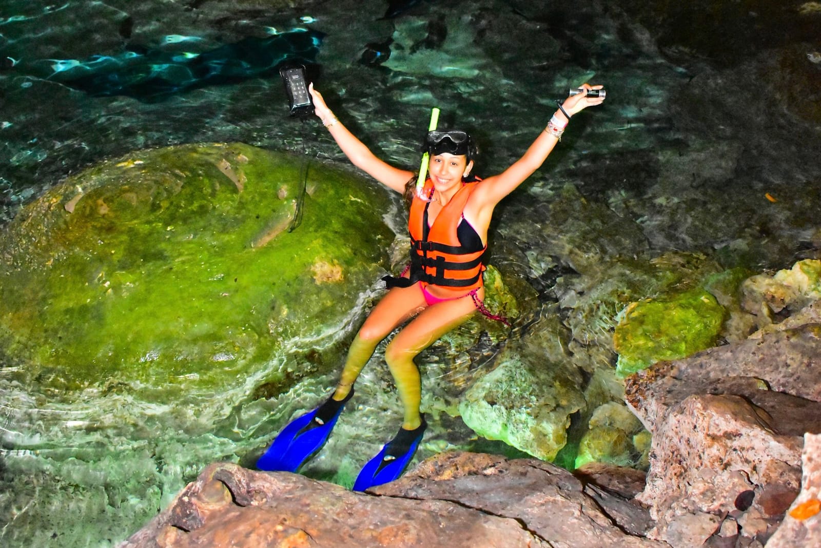 Visitante con aletas y chaleco celebrando sobre una roca del cenote, con su lámpara sumergible en la mano