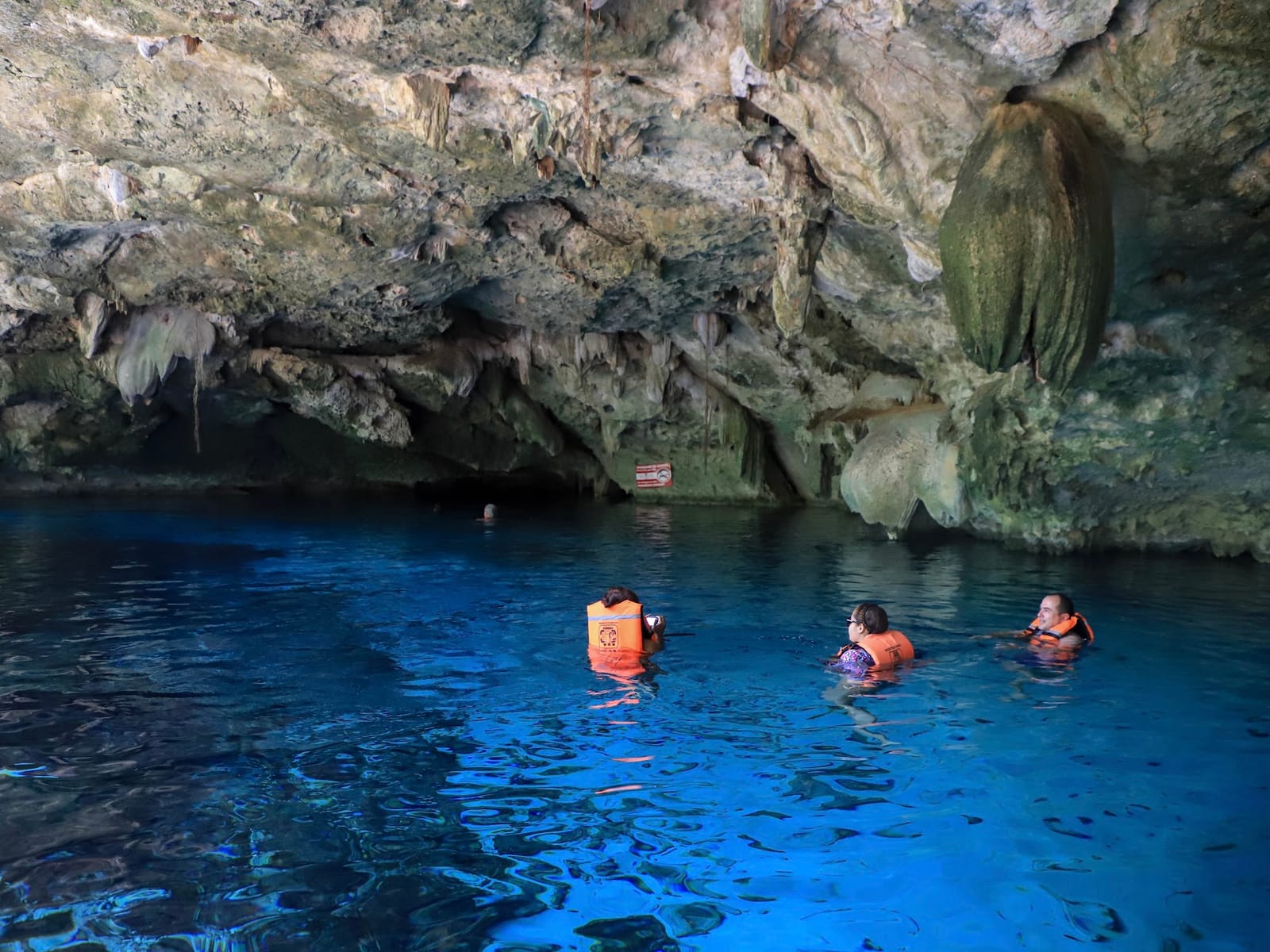 Tres visitantes con chaleco flotando bajo una gran estalactita en el Cenote Dos Ojos