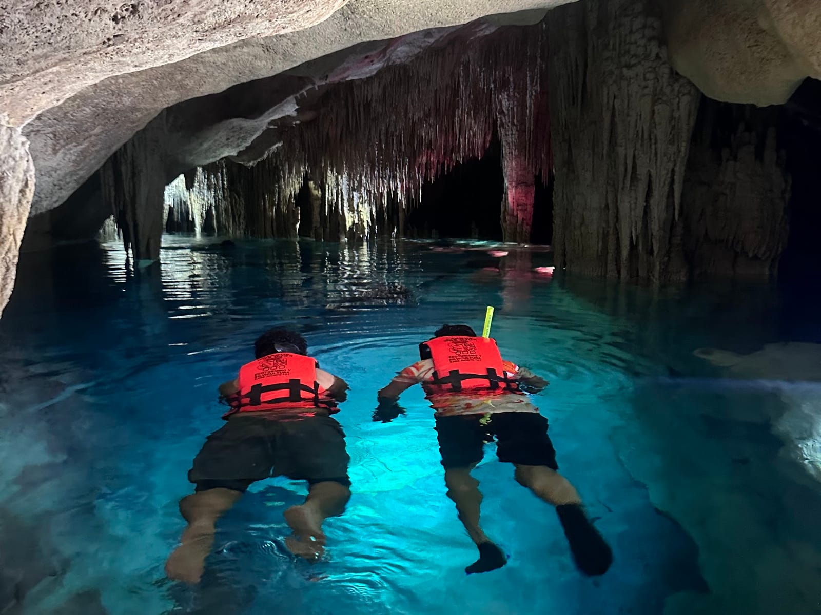 Zwei Schnorchler mit Schwimmweste gleiten durch einen beleuchteten unterirdischen Fluss zwischen Tropfsteinen, Cenote Los Monos