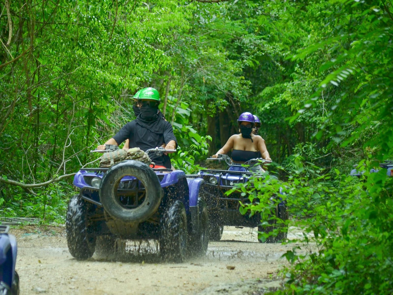 ATVs riding in line through a green jungle tunnel, helmets on and bandanas against the dust