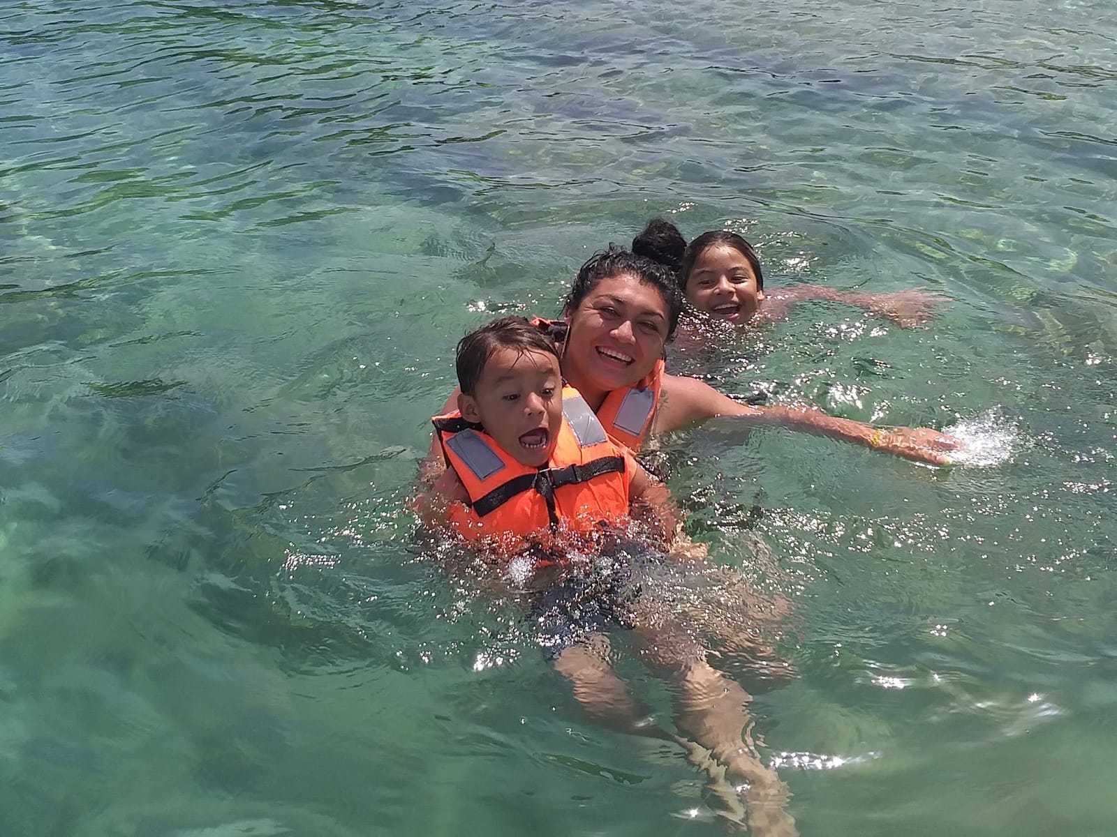Mamá y niños con chaleco salvavidas riendo mientras nadan en el agua verde y clara de la laguna