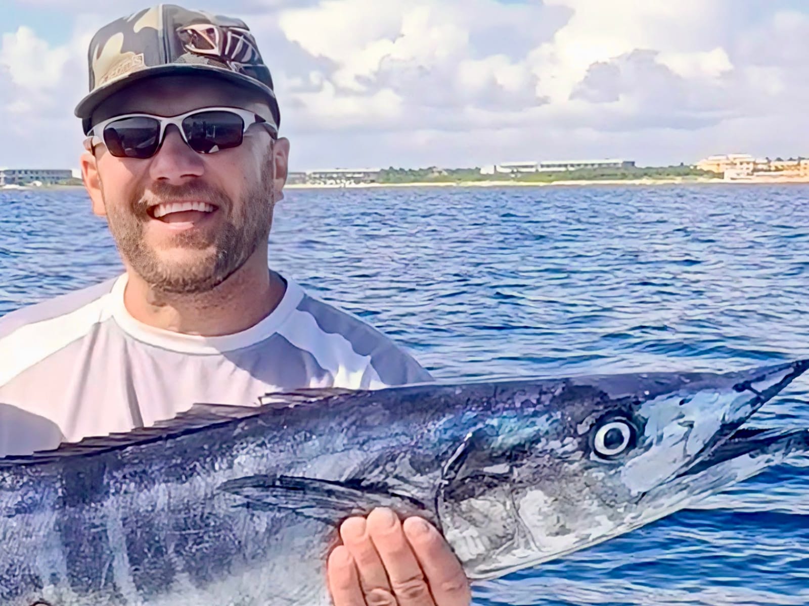 Pescador sonriente con un wahoo a bordo, con la costa al fondo