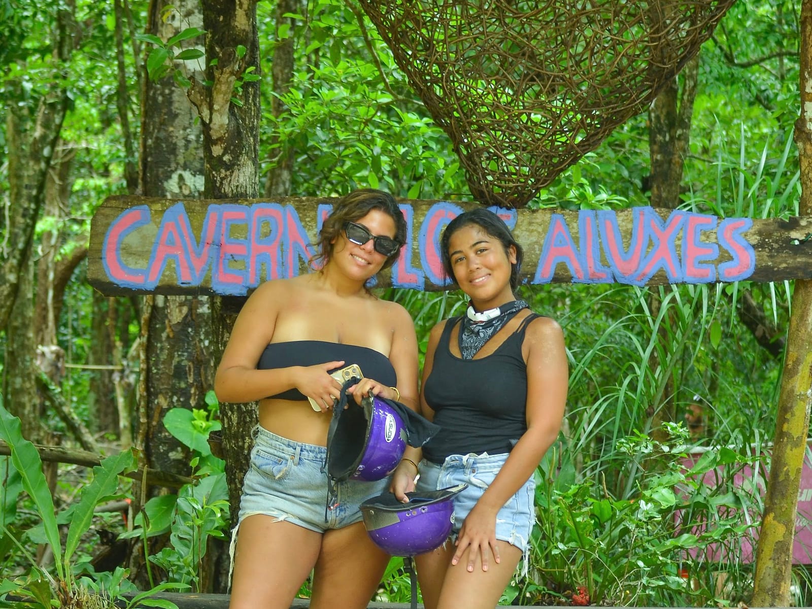 Dos amigas con sus cascos junto al letrero de la Caverna de los Aluxes